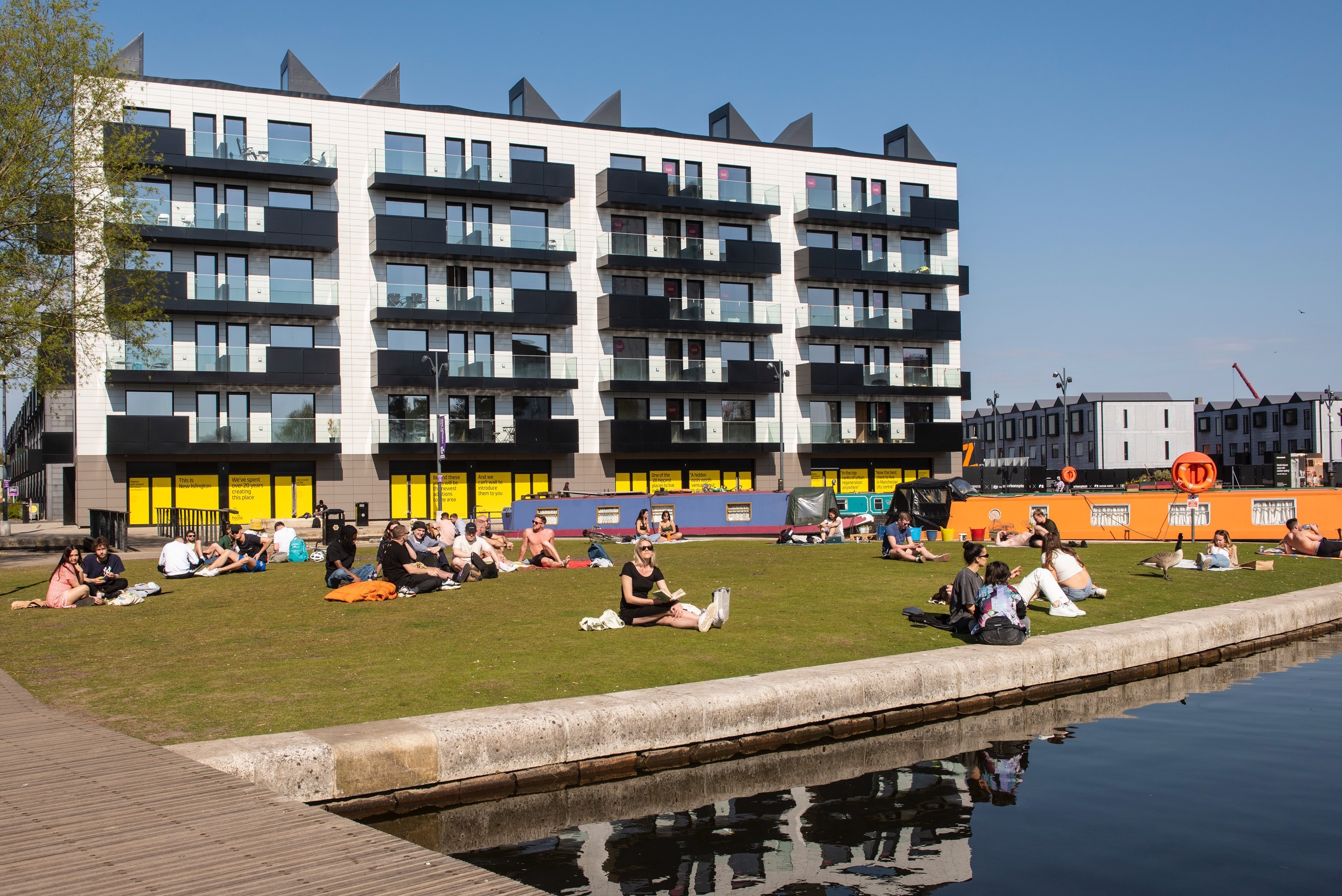 People relaxing on a grassy area next to a canal, with a modern apartment building in the background.
