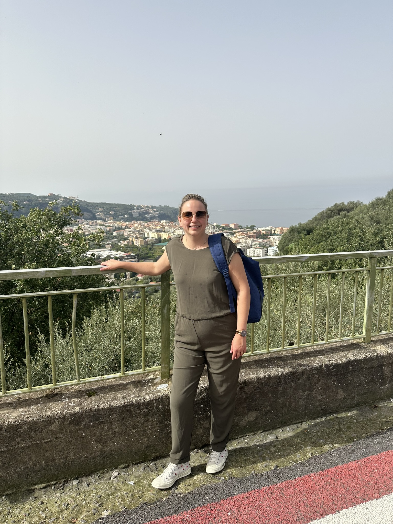 Woman in olive jumpsuit and sunglasses at overlook with coastal town in background.