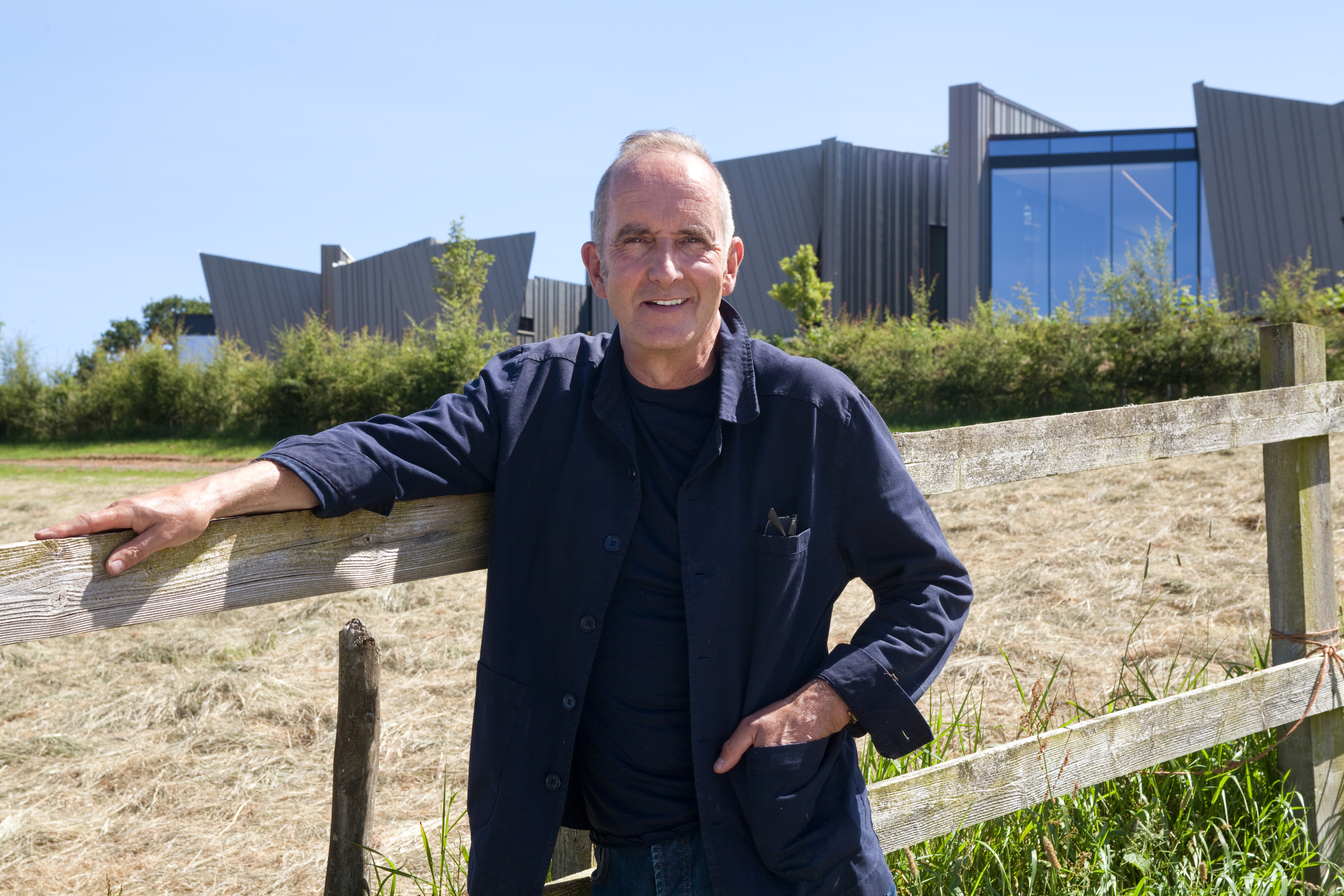 Kevin McCloud standing by a fence with a modern house in the background.